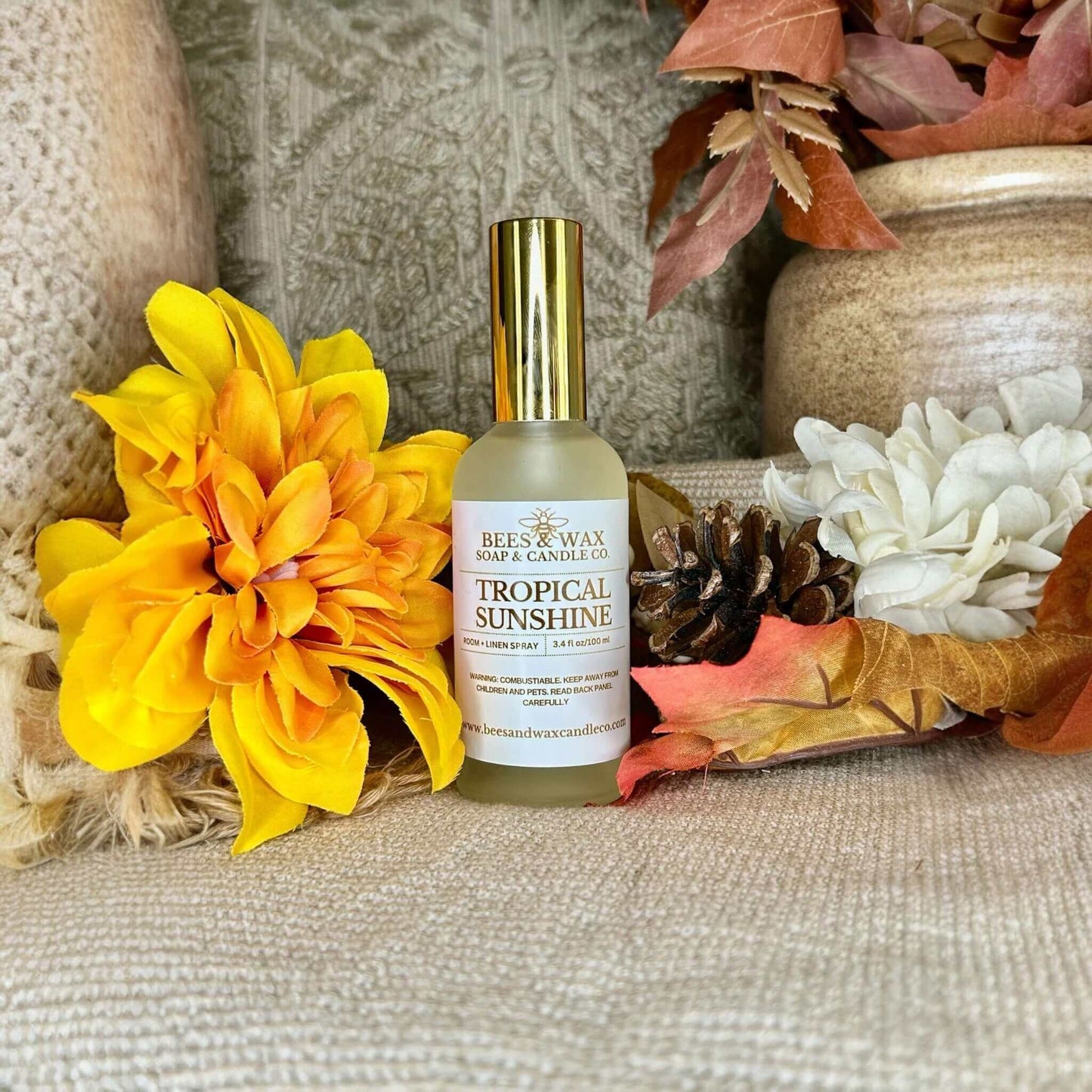 A bottle of tropical sunshine room + linen spray surrounded by vibrant flowers and cozy decor.